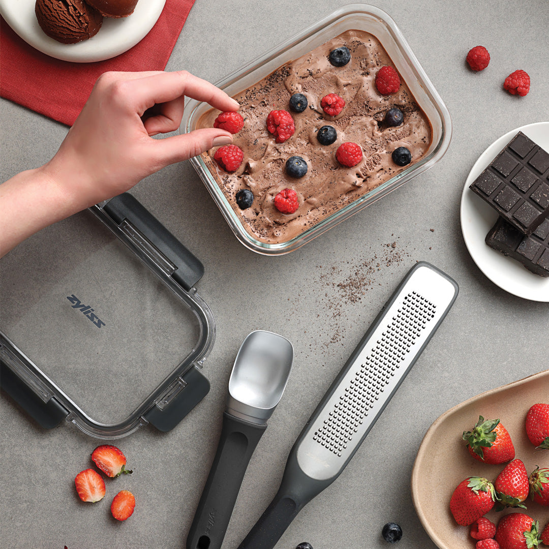 #belugagrey A hand decorates a container from the Zyliss Dessert Set with fresh berries. Beside it are a metal scoop, glass storage boxes, strawberries, chocolate pieces, and scattered berries on a gray countertop.