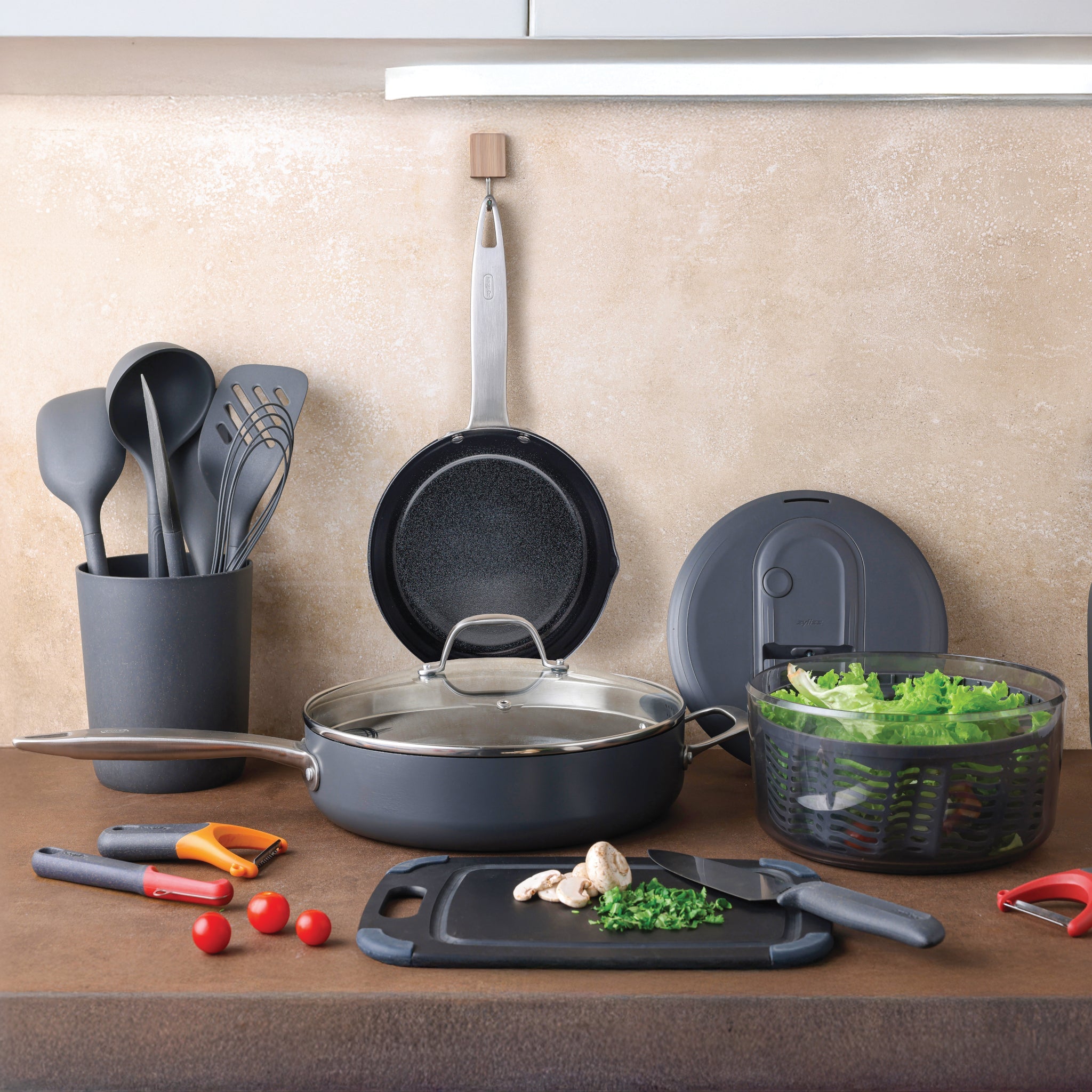 A kitchen countertop with various utensils, a cutting board with a knife, mushrooms, and chopped herbs, a skillet with lid, a hanging frying pan, salad spinner with greens, and several kitchen tools in a holder.