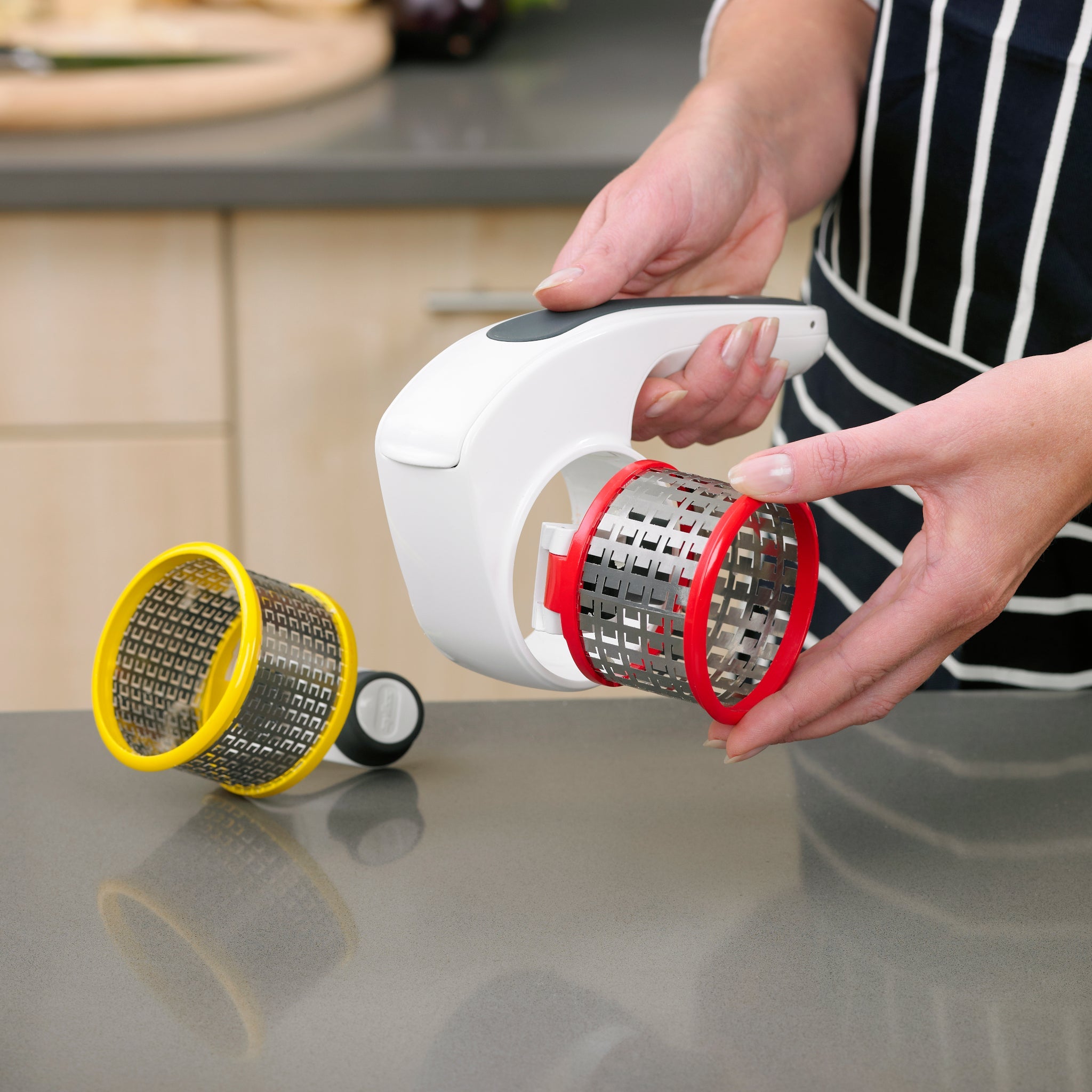 A person in a striped apron is assembling a handheld rotary grater with interchangeable drum blades on a kitchen counter; two grating drums, one with a yellow rim and one with a red rim, are visible.