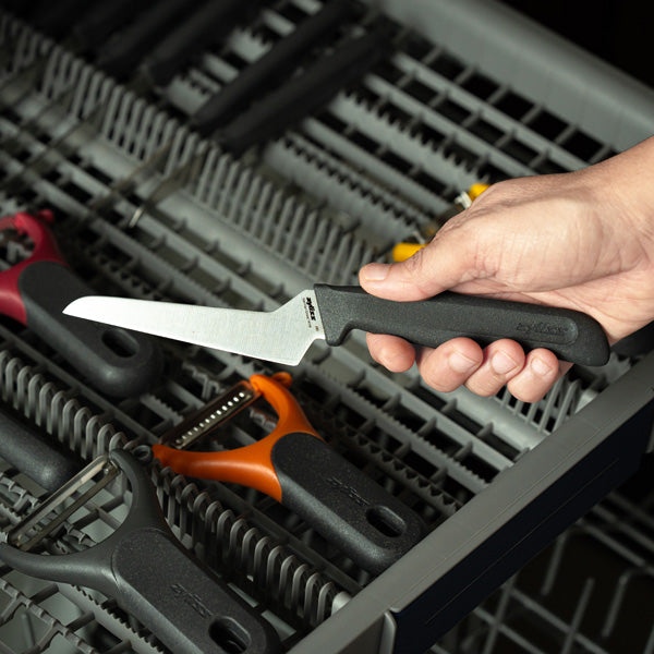 A hand places a kitchen knife with a black handle into a dishwasher utensil rack next to several vegetable peelers with black, red, and orange handles.
