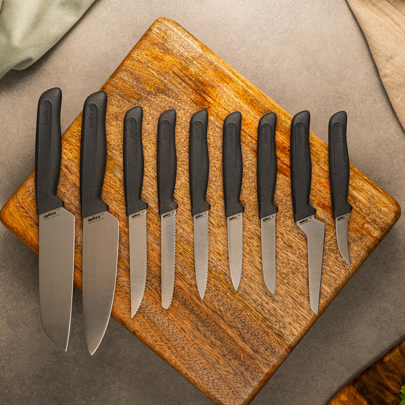 A set of eight kitchen knives with black handles arranged in a row on a wooden cutting board, displayed on a gray surface with cloths partially visible in the corners.