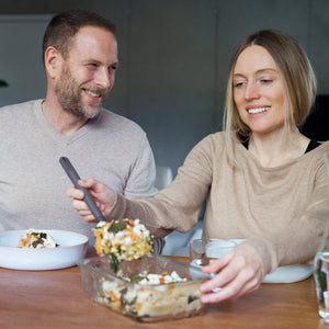 A smiling woman serves a casserole at a wooden table, with Zyliss Glass Containers nearby for easy meal prep. She and the man beside her look relaxed, enjoying a cozy indoor meal together. #0.37L/0.39qt