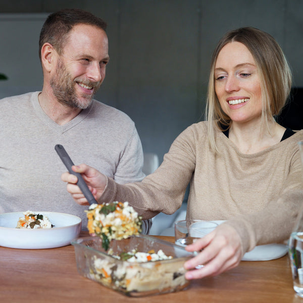 A smiling woman serves casserole from a Zyliss Glass Container with a secure seal as a man looks on. Both enjoy their meal at a wooden table, appreciating the convenience and easy prep offered by Zylisss storage solution. #0.64L/0.67qt