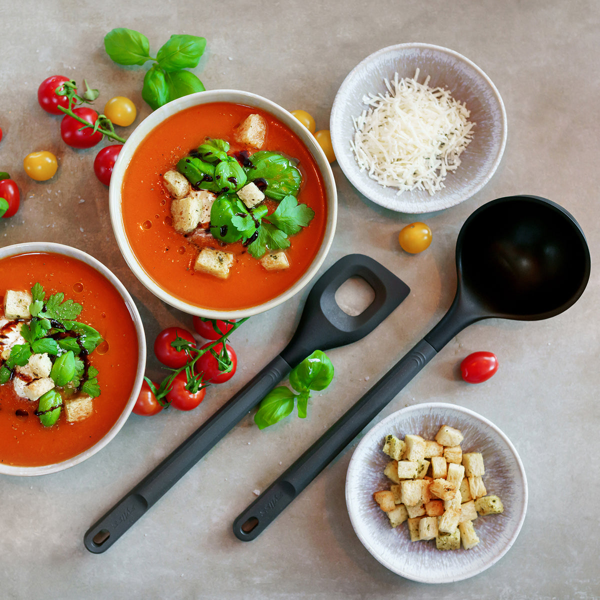 Two bowls of tomato soup topped with croutons and herbs, surrounded by fresh cherry tomatoes, a bowl of shredded cheese, a bowl of croutons, basil leaves, and two black soup ladles on a light surface.