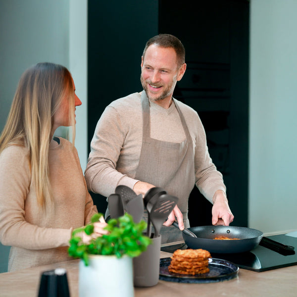 A man in an apron cooks at the stove with a Zyliss Spatula, smiling at a woman. Its silicone head meets heat-resistant needs as they stand in a modern kitchen with pancakes and a potted herb on the counter. #Small