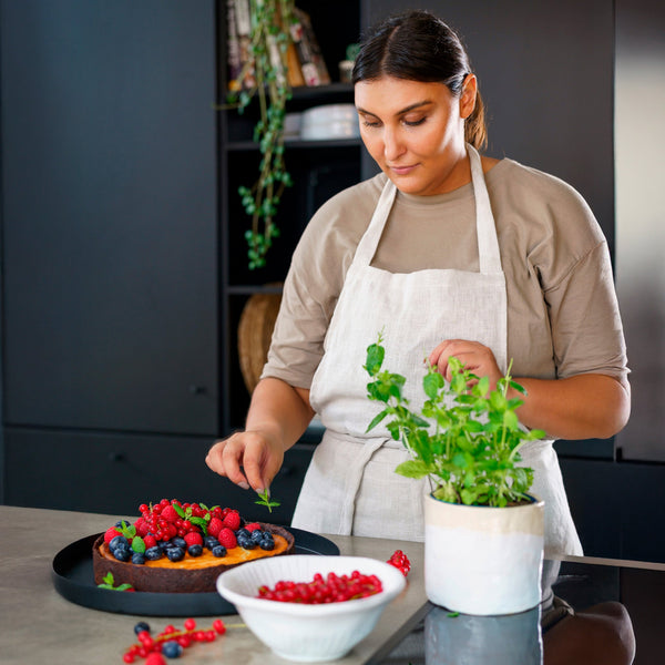 In a modern kitchen, a woman in an apron uses a Zyliss Spatula to add fresh mint leaves to a berry-topped cake. A bowl of berries and a potted herb plant are on the counter nearby. #Medium