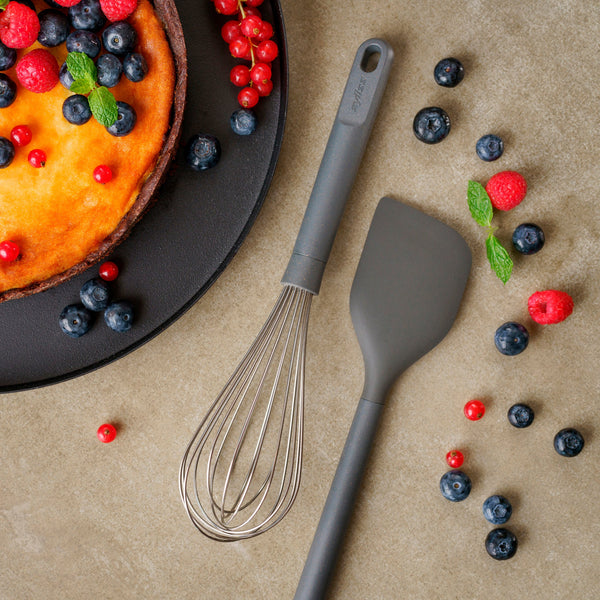 A cheesecake topped with berries sits beside a whisk and a Zyliss Spatula on the countertop, with blueberries, raspberries, and red currants scattered around. #Large